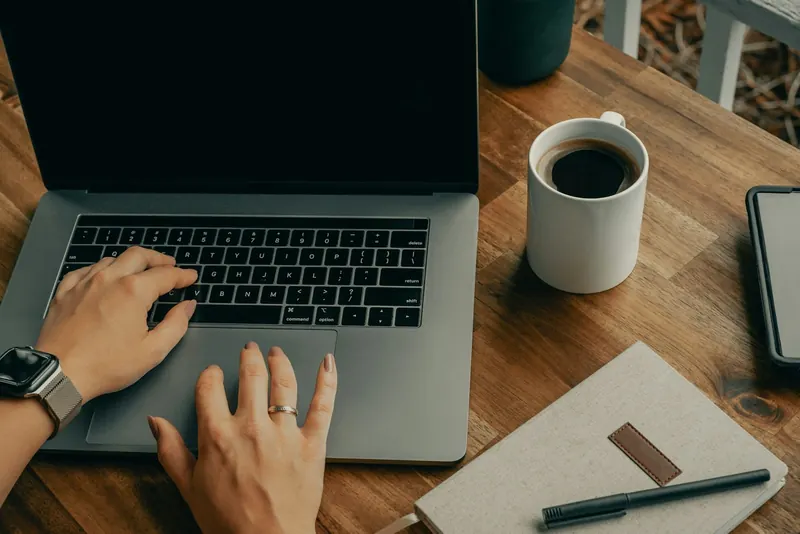 Hände auf einem Laptop, daneben Kaffeetasse und Notizbuch